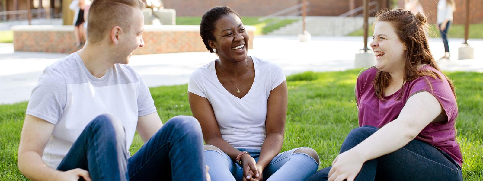 Two female students and one male student sitting on grass on forum laughing